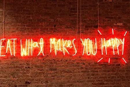 A neon sign on a brick wall that says eat what makes you happy
