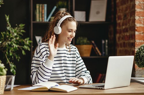 Cheerful-woman-in-headphones-greeting-friend-while-talking-on-laptop-at-home