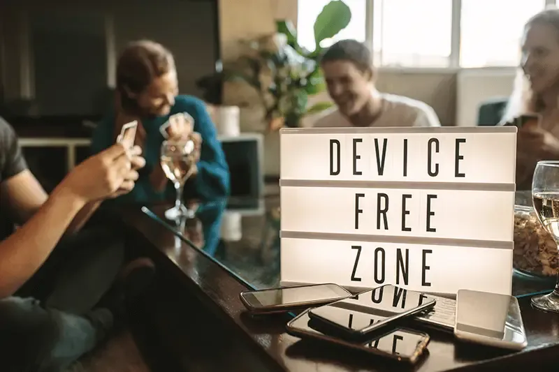 Friends hanging out laughing next to a Device Free Zone lightbox sign and a pile of mobile phones