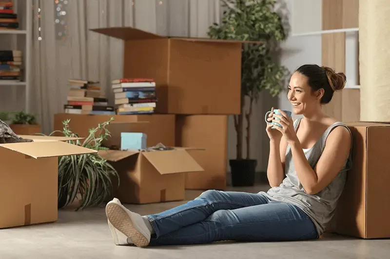 Happy woman resting and drinking coffee sitting next to moving boxes