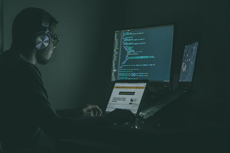 Man in a darkened room using numerous computer screens to hack others