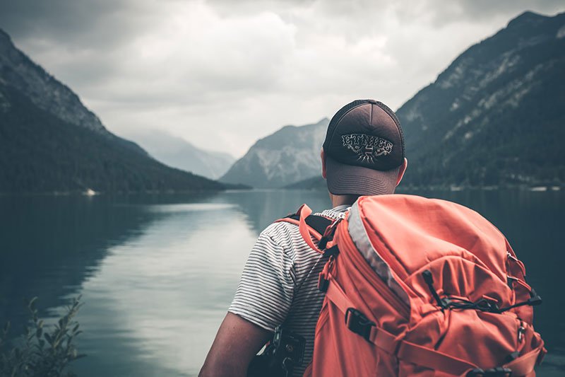 Man wearing a red hiking backpack and black backwards hat looking out at lake and mountains scenery