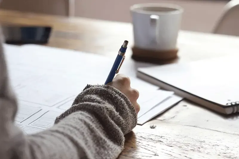 Person writing on paper on a brown wooden table with a notebook and white ceramic mug in the background