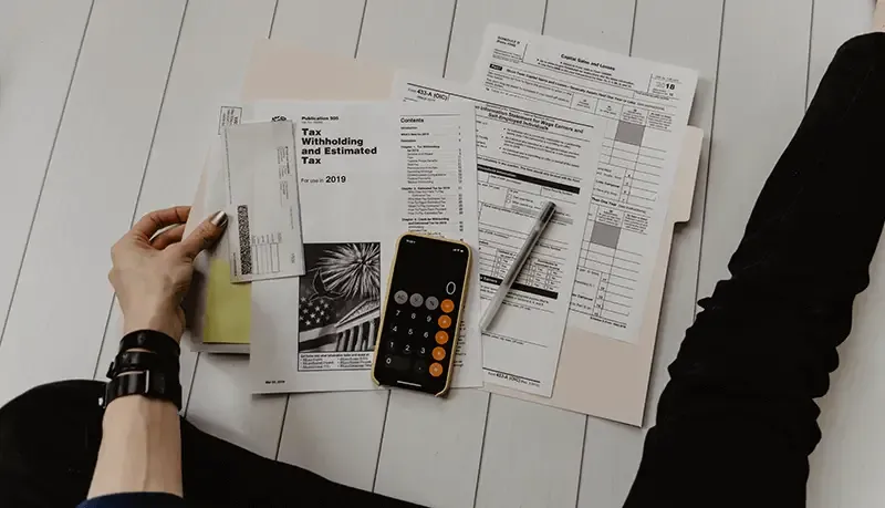 Person looking over their tax documents on the floor alongside a pen and calculator