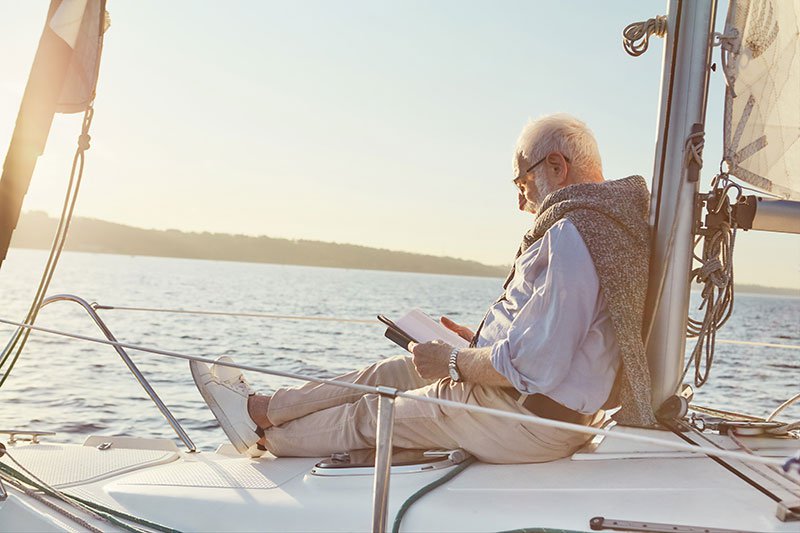 Relaxed senior man reading a book, while sitting on a sail boat deck floating in sea