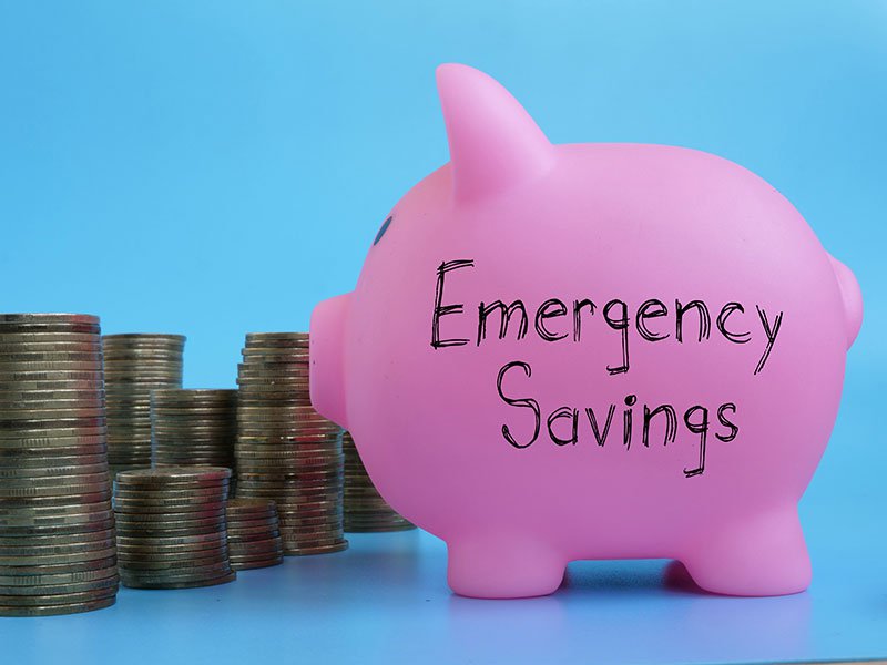 Stack of coins next to a pig money box with emergency savings written across it