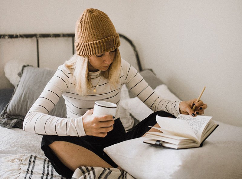 Woman in striped shirt sitting on bed writing and drinking coffee