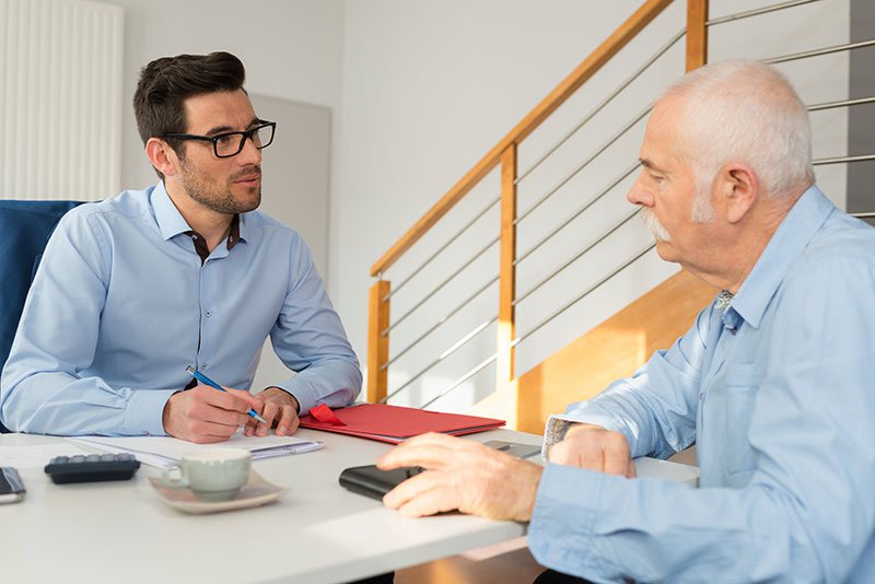 Young man speaking with older man and writing notes at desk