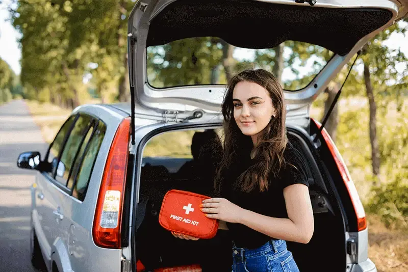 Young girl standing near open back door of silver hatchback car holding first aid kit