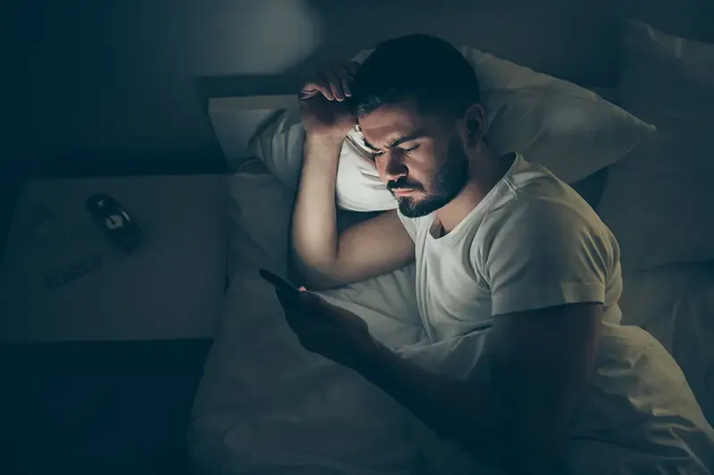 Guy lying in bed in the dark with insomnia, frowning and looking at his phone