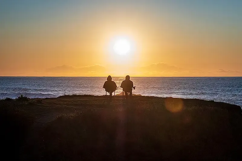 an elderly retired couple sitting on a beach watching the sun set