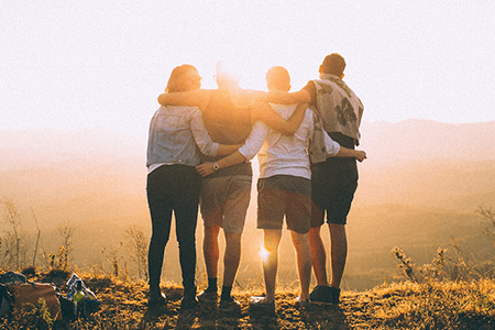 four-young-people-standing-on-hill-top-with-arms-around-each-other-gazing-at-the-sunset