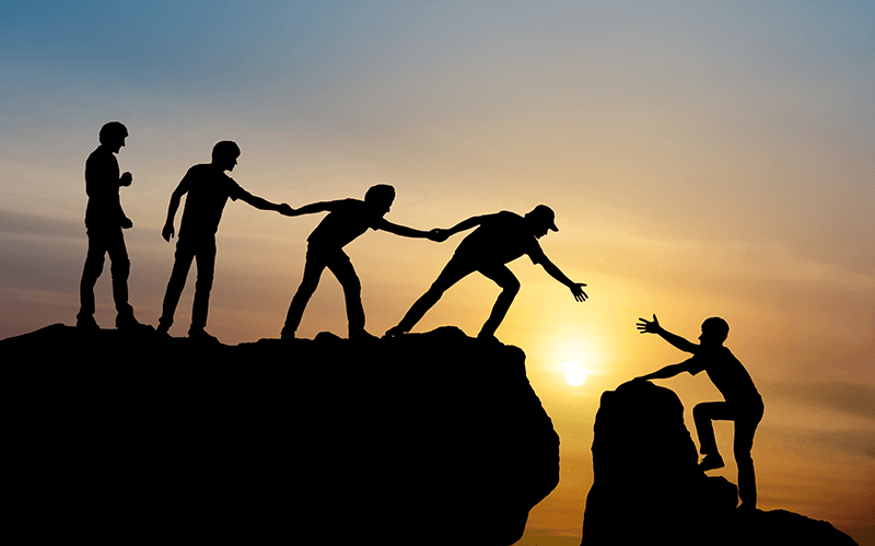 group-of-people-on-one-side-of-a-cliff-leaning-down-to-help-one-person-climb-the-other-side-of-the-cliff