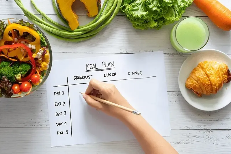 hand filling meal plan on weekly table with salad and fresh vegetable on dining table