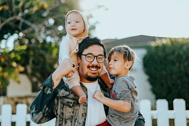 man in white tshirt and denim jacket holding a young boy around his waist and a little girl on his shoulders