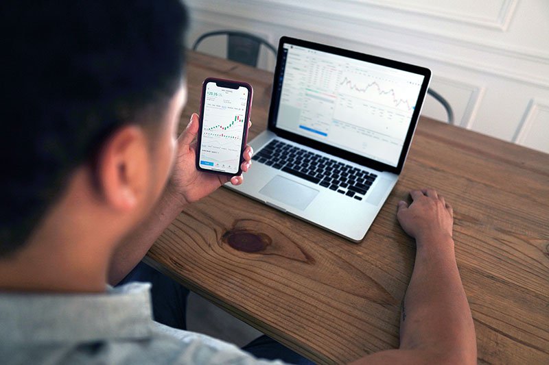 man looking at his investment portfolios on a laptop and his phone that is in his hand