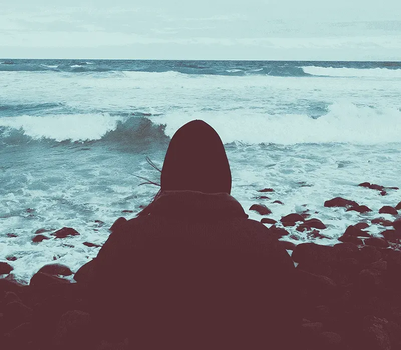 Person sitting on rocks rugged up and watching the rough ocean waves