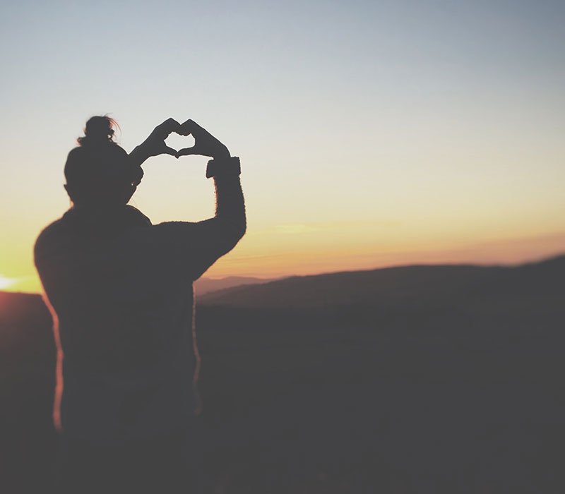 person-standing-on-clifftop-with-fingers-in-heart-shape-watching-the-sunset