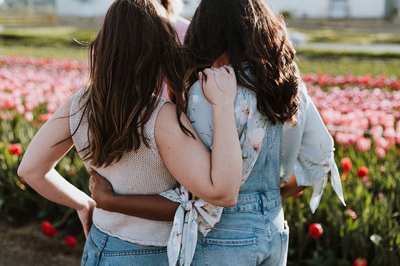two ladies with their arms around each others backs, stating into a flower field
