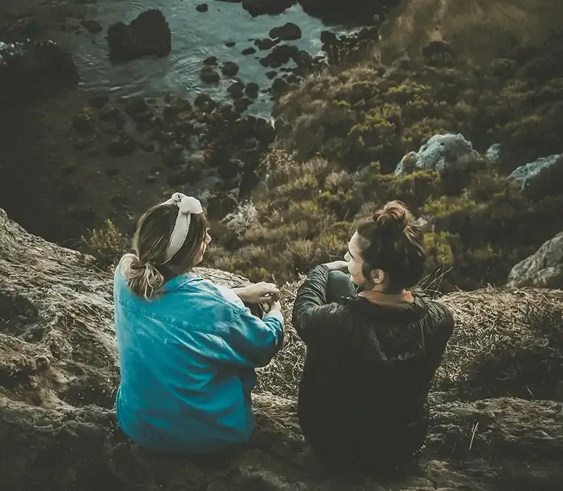 two women sitting and talking on a cliff edge with water and trees below