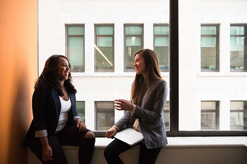 two women sitting on a window ledge smiling and laughing