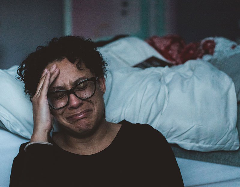 woman crying on floor leaning against a bed with her head in her hand