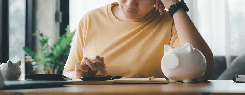 woman in yellow tshirt sitting at a table using a calculator next to a piggy bank