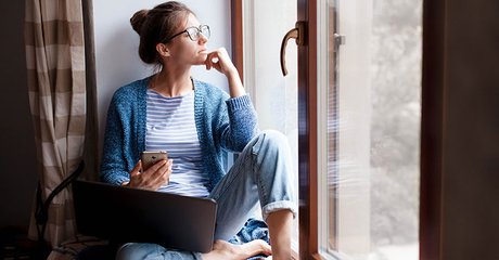 woman-thinking-and-looking-out-window-while-working-on-her-laptop-and-phone