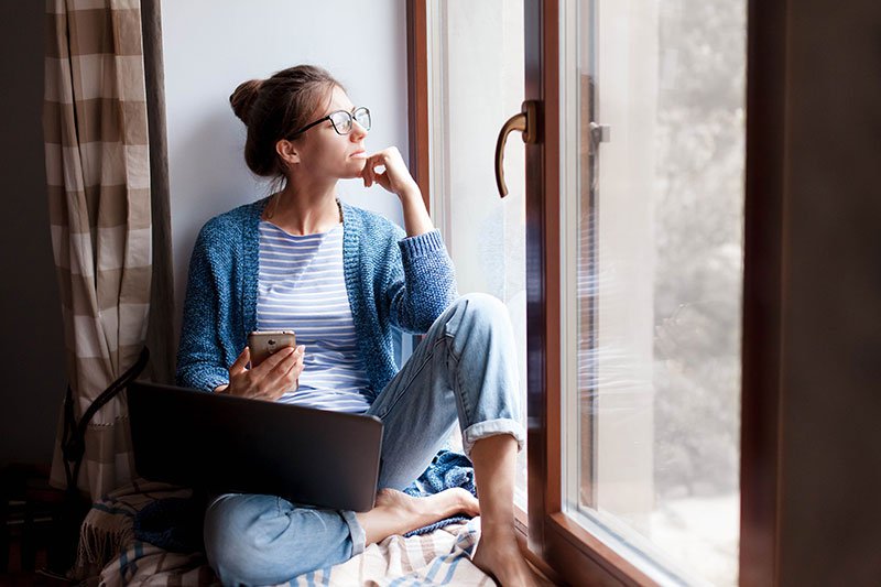 woman-thinking-and-looking-out-window-while-working-on-her-laptop-and-phone