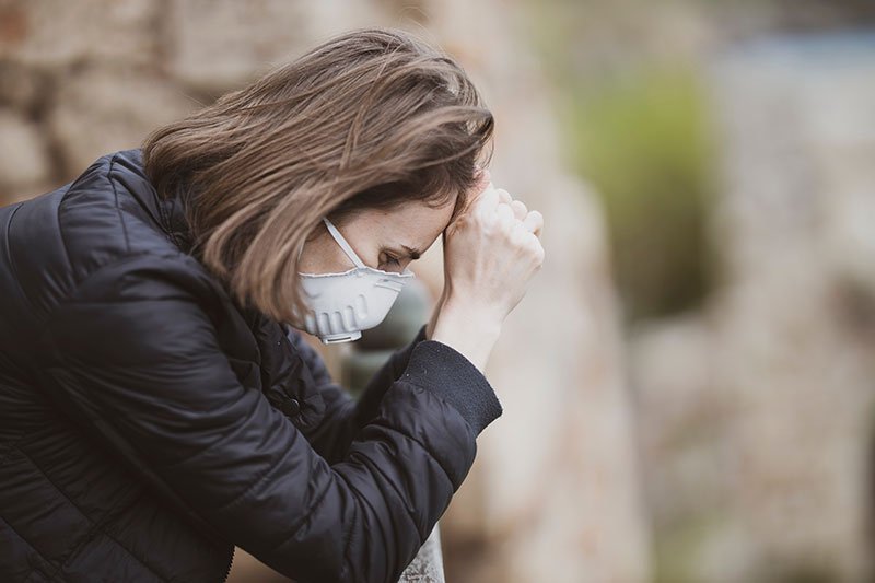 woman leaning against a fence with her head in her hands wearing a face mask