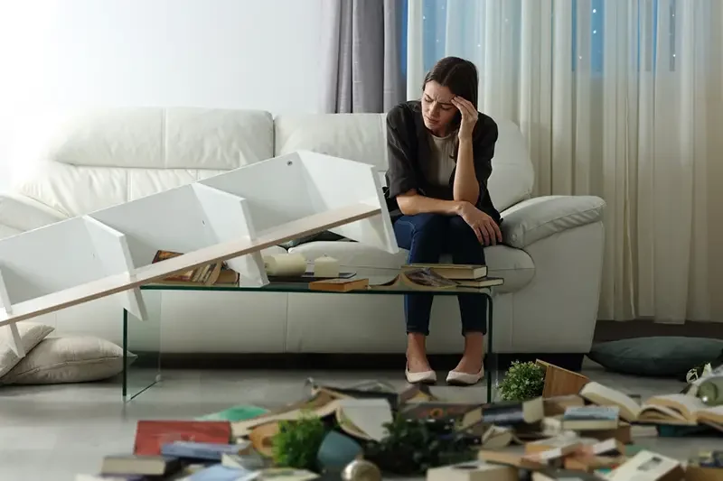 woman upset on couch with bookshelf fallen over and books everywhere