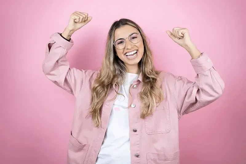 Young happy woman in a pink jacket and wearing glasses with her arms in air against a pink background